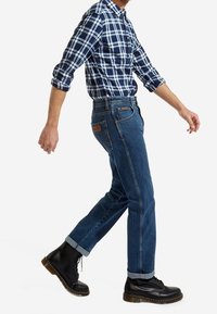 Vaqueros de mezclilla azul con un corte recto clásico, combinados con una camisa de cuadros en tonos azul marino y blanco. Botas negras de cordones con suelas texturizadas.