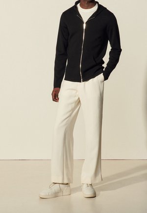 Homme portant un sweat à capuche noir zippé, un t-shirt blanc, un pantalon large crème et des baskets blanches, debout devant un fond clair.