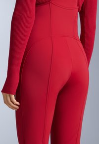 Piros testhez simuló overall bordázott hosszú ujjakkal, sima textúrával és varrási részletekkel. Magasított derékkal és formázott hátrész kialakítással rendelkezik.