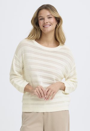 Ung kvinde med langt blondt hår iført en cremefarvet, stribet striktrøje og beige bukser, smilende med hænderne løst foldet sammen.