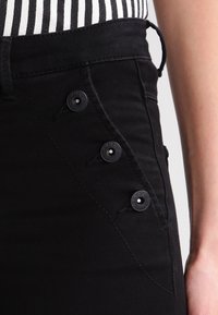 Černé džíny mají přiléhavý střih s vysokým pasem, pět černých knoflíků po straně a hladkou texturu. Zdůrazněny minimálním šitím.