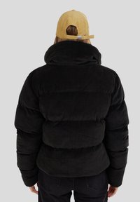 Mustapintainen corduroy-toppatakki, jossa on korkea kaulus ja suuret tikattuja osia. Mallilla on päällään keltainen cord-kappi, jossa on säädettävä takapuoli.