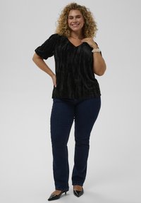 Lächelnde Frau mit lockigem Haar, die ein schwarzes Samtoberteil, dunkelblaue Jeans, schwarze High Heels, ein Perlenarmband und eine goldene Halskette trägt, posiert vor weißem Hintergrund.