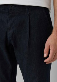 Pantaloni in denim scuro con taglio dritto, dotati di chiusura con bottone, tasche anteriori e una texture sottile. Il tessuto appare resistente e liscio.