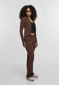 Barna, illeszkedő, cipzáros dzseki és hozzá illő nadrág puha, rugalmas anyagból; ujjtámlákkal és fekete, illeszkedő felsővel alatta.