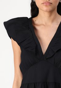 Vestido preto com decote em V profundo, detalhes em babados nos ombros e cintura franzida para uma silhueta fluida. Tecido suave e texturizado.