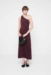 Femeie îmbrăcată într-o rochie midi burgundy cu o singură umăr, ținând o geantă neagră și stând în pantofi cu toc ascuțit negri, cu fundițe.