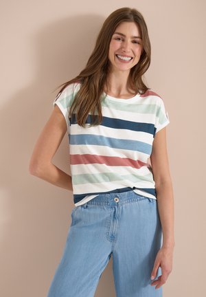 Femme souriante, vêtue d’un T-shirt à manches courtes avec des rayures horizontales multicolores et d’un pantalon bleu clair à taille élastique, debout contre un mur beige.