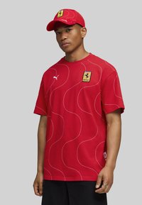 Mand iført en rød Puma Ferrari t-shirt med bølgede hvide linjer og en matchende rød Ferrari kasket, stående mod en ensfarvet grå baggrund.