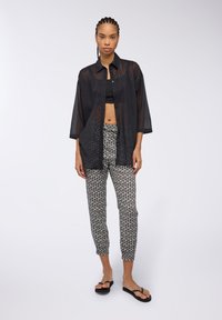 Camicia nera trasparente con bottoni indossata sopra un top nero corto, abbinata a pantaloni bianchi a righe con gamba affusolata e infradito neri.