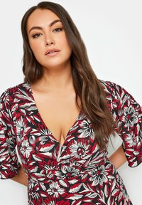 Rotes, weißes und schwarzes Blumenkleid mit kurzen Ärmeln und einem tiefen V-Ausschnitt. Der Stoff zeigt ein kräftiges Blumenmuster mit strukturierten Details.
