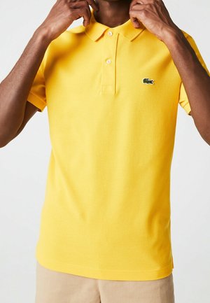 Man adjusting collar of bright yellow polo shirt with small green crocodile logo, beige pants visible at bottom.