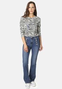 Mujer de pie vistiendo una camiseta de manga larga con estampado de cebra, jeans de tiro alto azul con botones en el frente y zapatillas blancas.