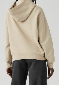 Felpa oversize beige con cappuccio regolabile con cordoncino, tessuto morbido e polsini a coste, abbinata a pantaloni in denim grigio scuro. Vista posteriore.