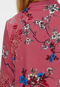 Tissu à motif floral en rose, présentant des fleurs blanches, bleues et rouges avec des détails linéaires complexes. Texture douce, encolure ronde.