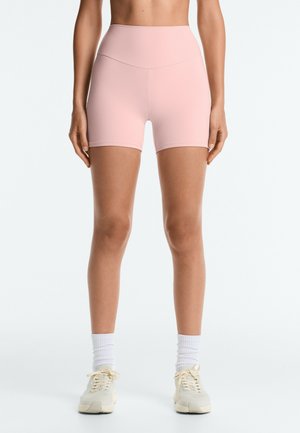 Femme portant un short cintré taille haute rose clair, des chaussettes blanches et des baskets beiges, debout avec les bras détendus le long du corps.