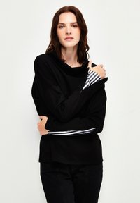 Maglietta nera a maniche lunghe con collo a cowl e tessuto a righe a contrasto sulle maniche. Texture morbida, vestibilità rilassata e dettagli di cucitura visibili.