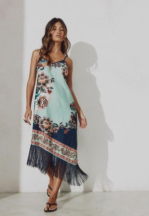 Mujer con un vestido sin mangas con estampado floral y flecos en el dobladillo, y sandalias negras, de pie junto a una pared clara que proyecta una sombra.