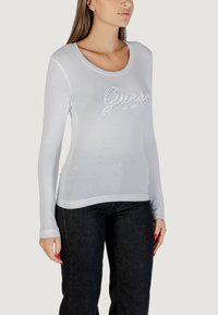 Blusa blanca de Manga Larga con escote redondeado, con un logo "Guess" texturizado embellecido con cristales, combinada con pantalones de mezclilla oscuros.