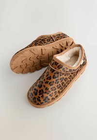 Zapatos tipo slip-on con estampado de leopardo, forrados con un pelaje sintético de color topo y suela de goma. Presentan una forma redondeada y adornos cosidos a lo largo de la parte superior.