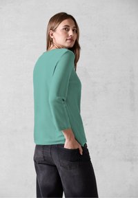 Türkises Langarmtop mit strukturiertem Finish, Rundhalsausschnitt und lässiger Passform. Kombiniert mit dunkler Jeans, seitlich betrachtet vor einem grauen Hintergrund.