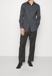 Modrá košile s dlouhým rukávem a bílým geometrickým vzorem, s knoflíkovým límečkem a rovný fit, v kombinaci s tmavými kalhotami a černými teniskami.