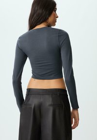 Frau mit langen dunklen Haaren, die ein enganliegendes, dunkelgraues langärmliges Crop-Top und hochtaillierte schwarze Lederhosen trägt, die von hinten zu sehen ist.