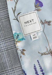 Étiquette de vêtement indiquant « NEXT TAILORING 40R Made in Vietnam » cousue sur une doublure en tissu fleuri à l’intérieur d’un vêtement gris à carreaux.
