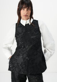 Sorte blomstret ermeløs topp med teksturert overflate, lagd over en hvit skjorte med krage. Antrekket har en asymmetrisk kant.