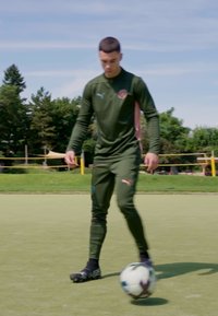 Sportos alkatú személy sötétzöld, testhez simuló melegítőben, pink részletekkel, egy mezőn állva, készülve egy fekete-fehér focilabda rúgására.