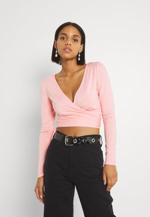 Top envolvente rosa claro de manga longa com decote em V e detalhe franzido, combinado com calças pretas de cintura alta e um cinto preto brilhante.