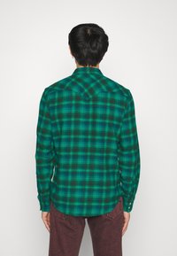 Camisa de cuadros verde con mangas largas y cuello puntiagudo. Está hecha de una tela tejida, con patrones de cuadros en negro y azul. Puños abotonados.