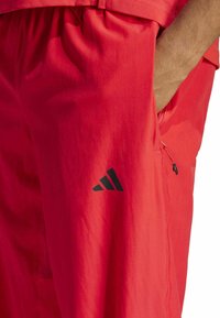 Red athletic pants with a smooth texture, featuring a side pocket with a zipper and a black Adidas logo on the lower leg.