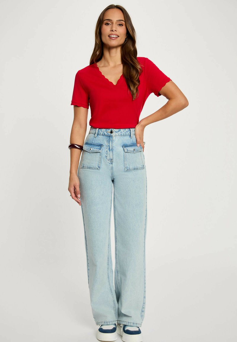 Donna con lunghi capelli castani che indossa una maglietta rossa a maniche corte e jeans a vita alta a gamba larga azzurri, in piedi con una mano sul fianco