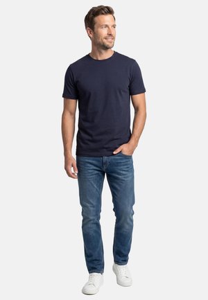 Mand står med hånden i lommen iført navyblå t-shirt, blå jeans og hvide sneakers på hvid baggrund.