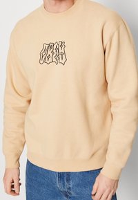 Mikina ze směsi bavlny v světlém béžovém provedení s černým abstraktním logem na hrudi. Disponuje kulatým výstřihem a žebrovanými manžetami.