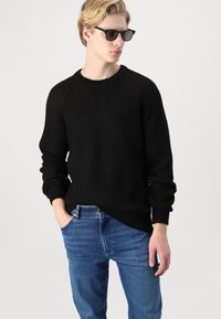 Czarny sweter z prążkowanej dzianiny z długimi rękawami, okrągłym dekoltem i fakturowanym splocie. W połączeniu z niebieskimi dżinsami. Gładkie wykończenie materiału.