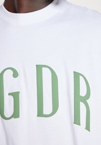 Vit bomull t-shirt med fet gröna blockbokstäver "GDR" över bröstet. Slät textur med klassisk rund halsringning.