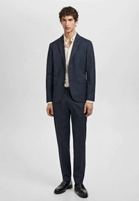 Costum în carouri bleumarin, cu o croială ajustată, sacou cu un singur rând de nasturi și pantaloni asortați. Purtați peste o cămașă de culoare deschisă, asociați cu pantofi negri.