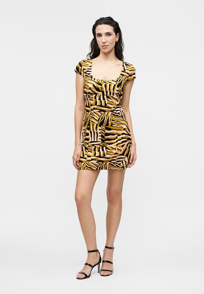 Γυναίκα φοράει κοντό, εφαρμοστό φόρεμα με κίτρινο, μαύρο και μπεζ αφηρημένο ζέβρα σχέδιο και μαύρα πέδιλα με λουράκια και ψηλό τακούνι.