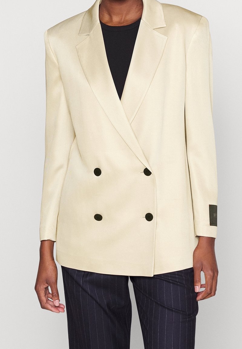 Blazer beige de doble botonadura con botones redondos negros, solapas en pico y textura suave, combinado con pantalones oscuros de rayas finas.