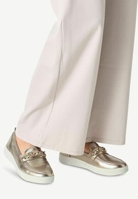 Chaussures à enfiler en métal doré avec une texture lisse, dotées d'un accent en chaîne et d'une semelle blanche, associées à un pantalon en tissu léger.