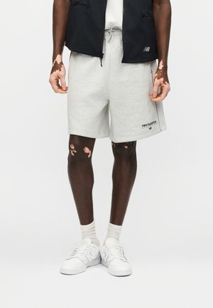 Model męski ubrany w jasnoszortowe spodenki New Balance, białe trampki, białe skarpetki i czarną kamizelkę na zamek, stojący na białym tle.