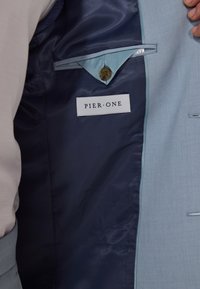 Parte interior de una chaqueta de traje azul claro con forro azul oscuro, cuenta con una etiqueta que dice "PIER ONE" y un botón cerca de un bolsillo. Textura suave.