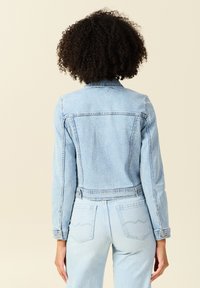 Chaqueta de mezclilla azul claro con un diseño corto, puños abotonados y costuras en el yugo trasero, que presenta dos bolsillos traseros con costuras curvas.