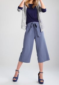 Ljusgrå capribyxor med knytmidja, kombinerade med en marinblå topp och en mönstrad vit cardigan. Blå platåsandaler fulländar looken.