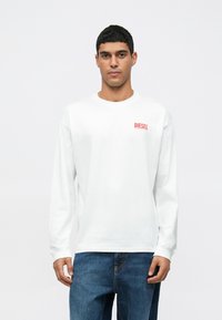 Camiseta de manga longa branca com um logo vermelho "DIESEL" no lado esquerdo do peito, combinada com jeans de denim azul. Textura lisa, design de gola redonda.