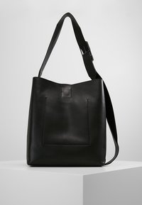 Czarna skórzana torba na ramię z szerokim, regulowanym paskiem i przednią kieszenią z przeszyciem, stojąca na białym postumencie na szarym tle.