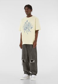 Žluté oversized tričko s květinovou modrou grafikou, spárované s volnými šedými džínami s dírami a bílými teniskami. Ležérní streetwear design.