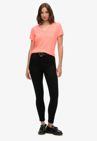 Neonroze V-hals t-shirt met korte mouwen, gecombineerd met zwarte skinny jeans en zwarte schoenen. Het shirt heeft een subtiele logo op de borst.